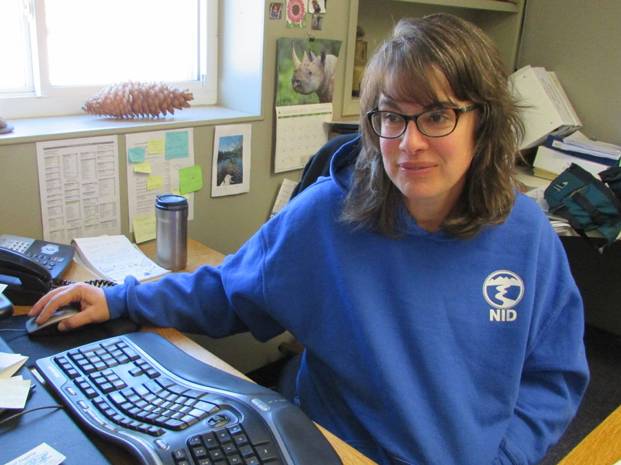 A woman with dark hair and glasses, wearing a blue sweatshirt, works at a computer on her desk.