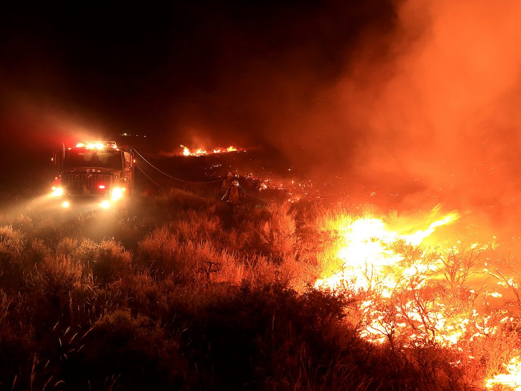 A wildfire burns hot in the Idaho sage brush as several fire fighters spray water hoses from a truck.