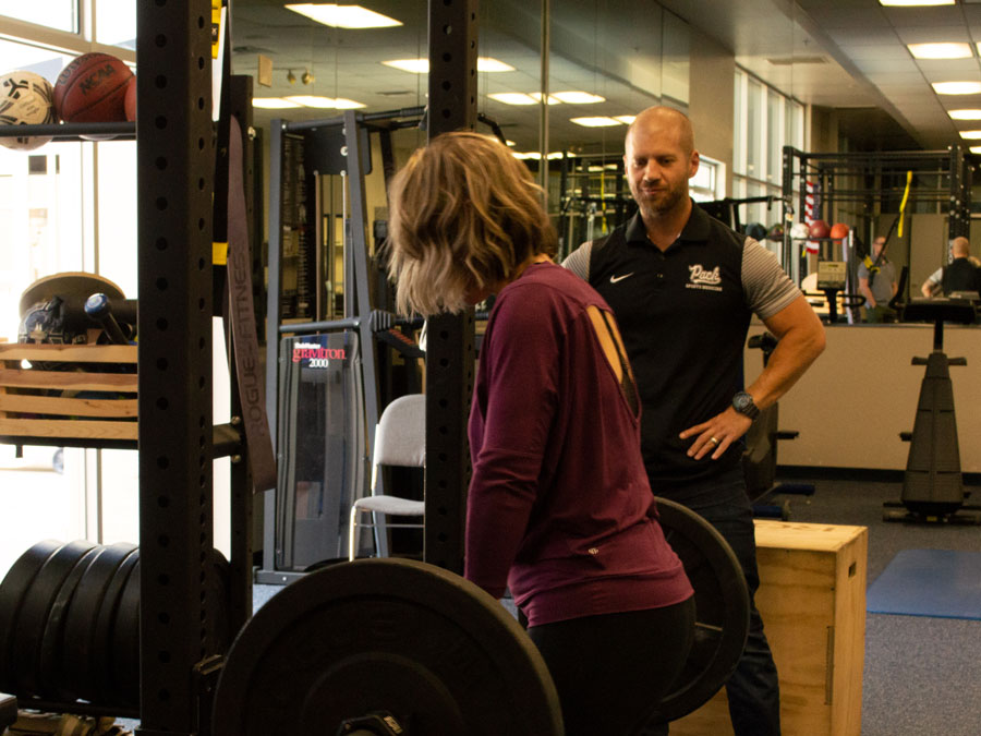 White man in a dark gym suit stands by a blonde woman lifting a weighted barbell.