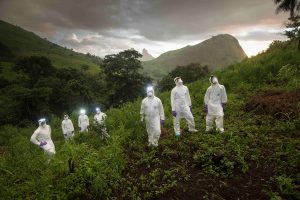 Researchers wearing all-white protective gear pose on a green hillside in Sierra Leone.