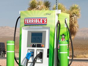 A chartreuse-green box-shaped charging station with two cords. The sign reads "terrible's." Yucca trees in background.