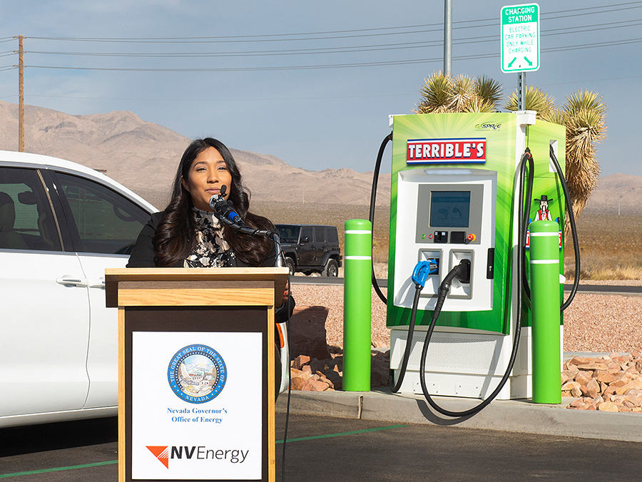 A brown-skinned woman with long black hair stands behind a podium that reads NV Energy. Behind her, a green EV charging station with two cords reads Terrible's.
