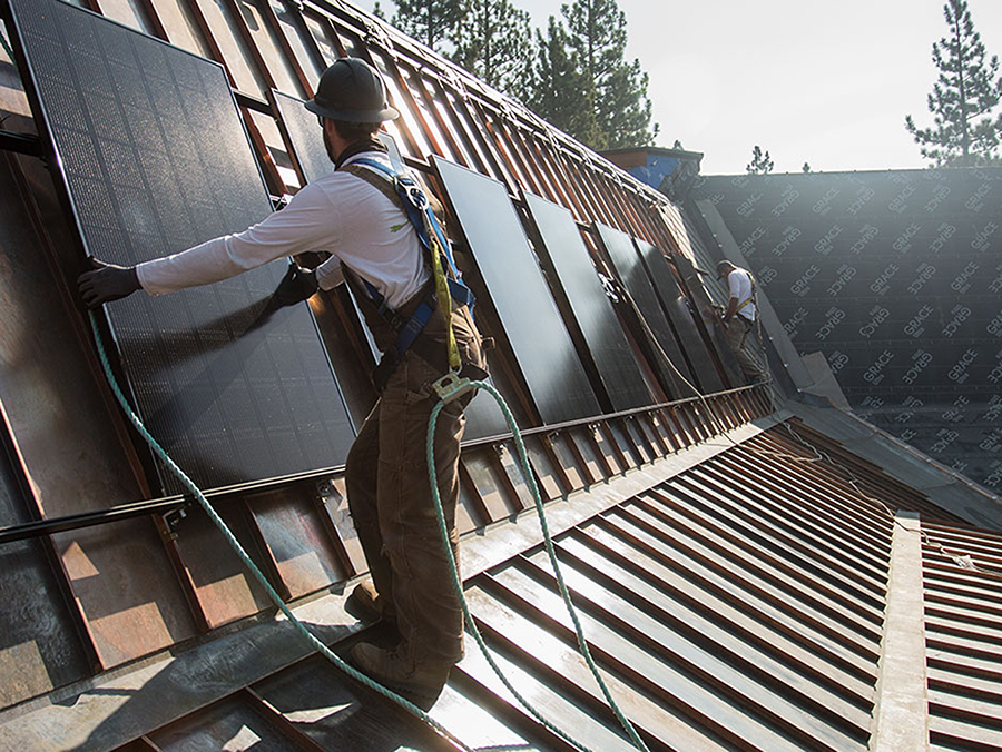 Two men standing on a brown mansard roof, insttalling shiny, rectangular panels.