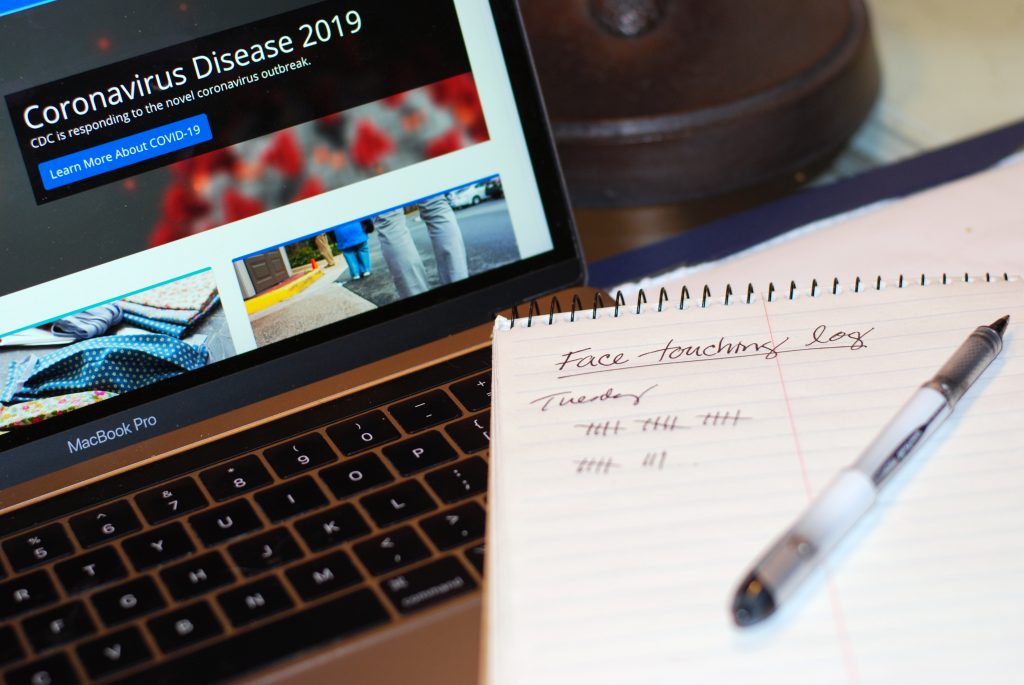 A pad of paper with a pen situated on it displays log of "Face Touching". A laptop screen is displayed in the background with information on the Coronavirus Disease 2019.