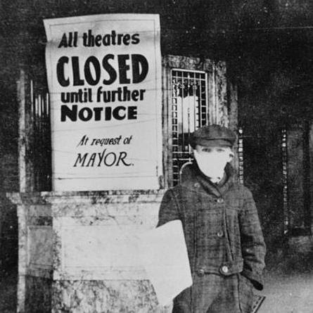 A person wearing a surgical mask stands next to a sign stating, "All theatres closed until further notice at request of mayor"