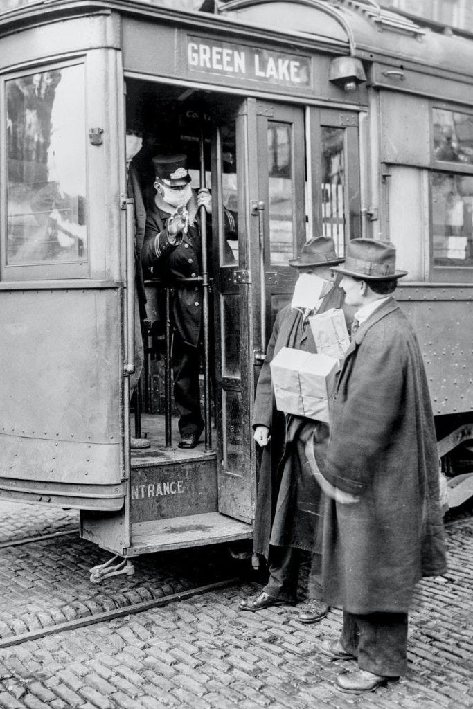 A train conductor wearing a mask speaks with two men - one of whom is wearing a mask, the other is not.