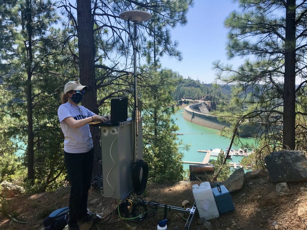 Ava Cooper works on a large piece of equipment with a dam and reservoir in the background.