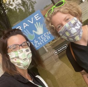 Two women stand with masks on in front of a RAVE sign