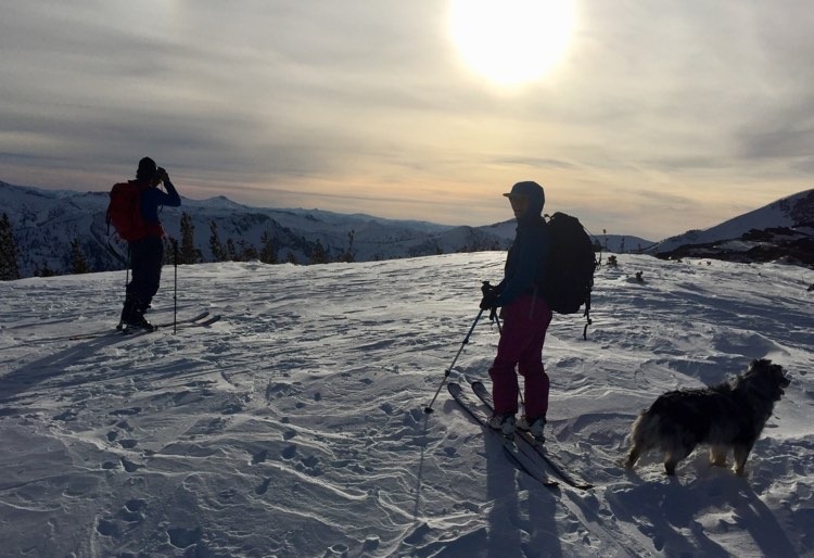 Two skiers and a dog stand in front of a Montana mountain landscape