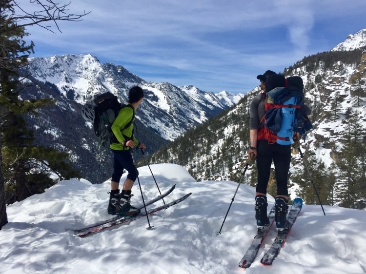 Two skiers with skins on their skis stand in front of a Montana mountain landscape
