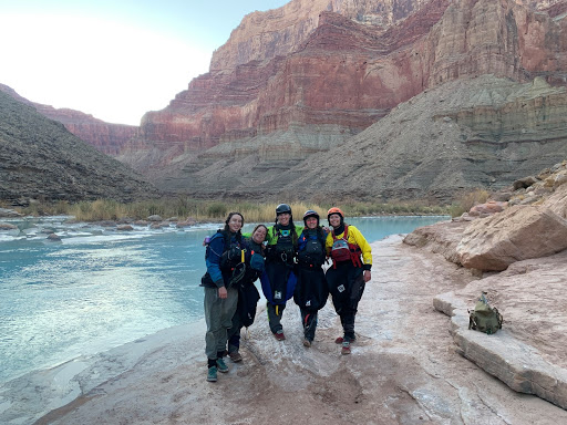 Five women in kayaking gear pose in front of a blue river, with a big canyon wall in the background
