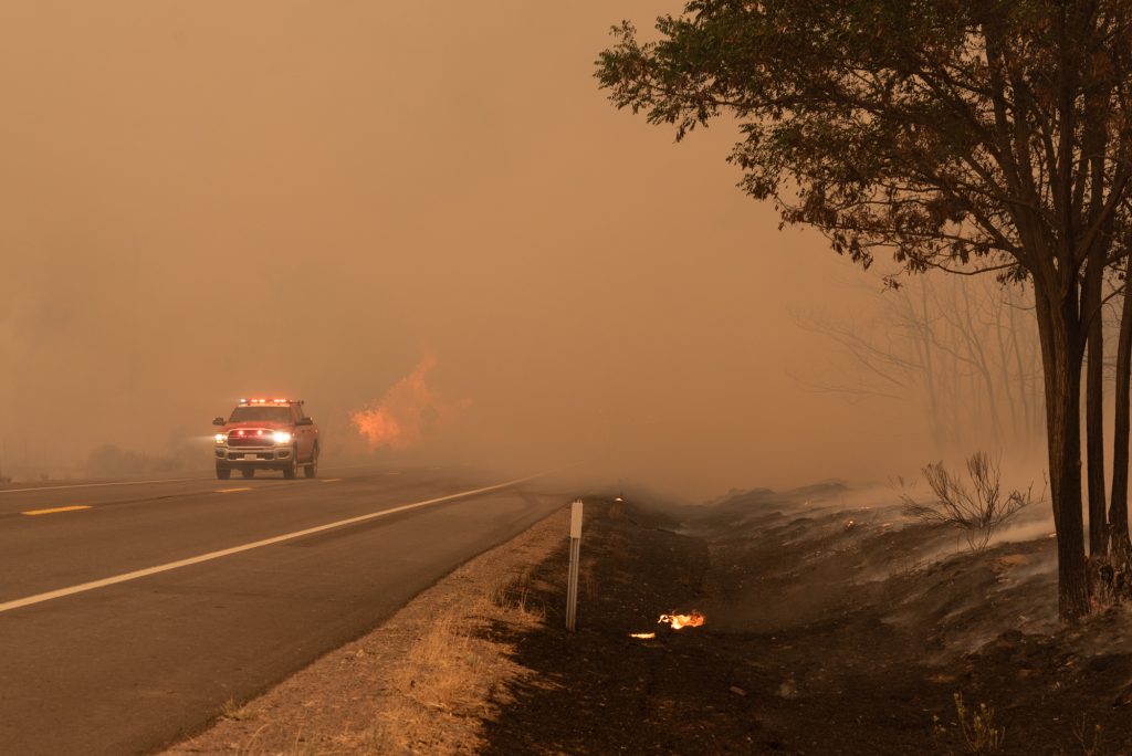 A red firetruck with lights on drives out of smoke with flames burning in the background.