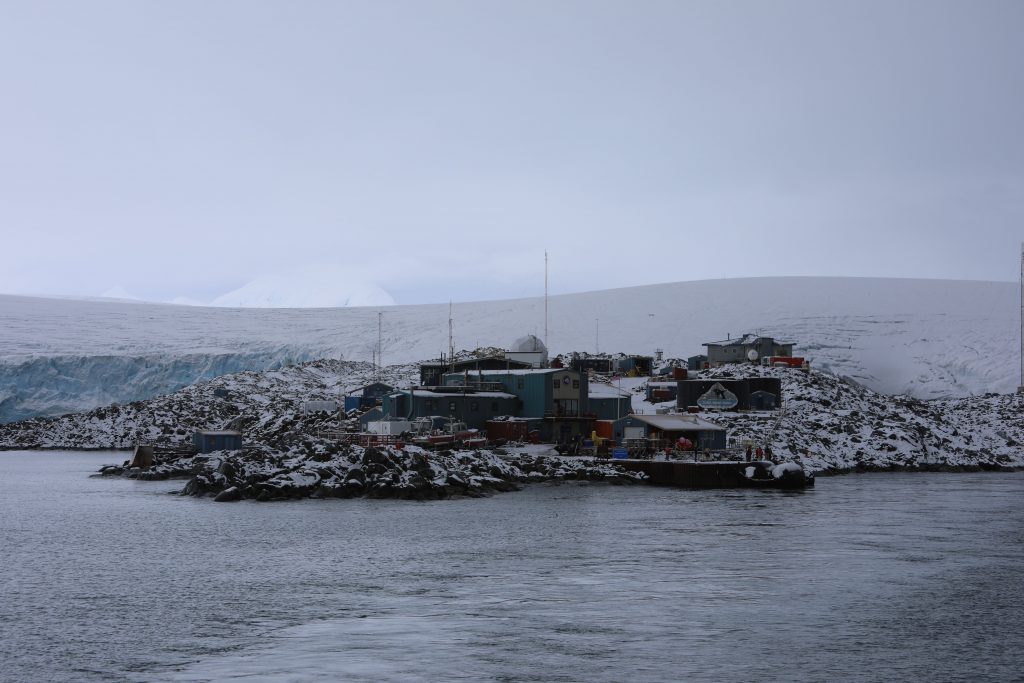 In the middle-ground, a frozen research station between the Antarctic Ocean in the foreground and the icy tundra in the background. It's comprised of some small, one-story metal buildings and a series of radio towers.
