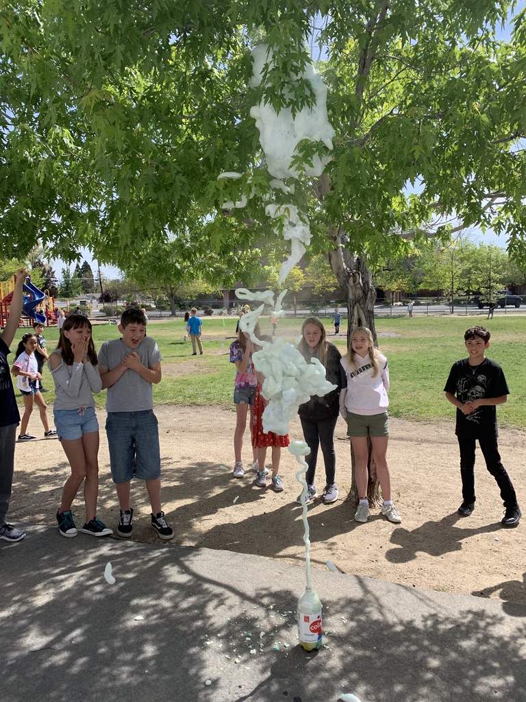 elementary-aged students gasp in wonder at some white-colored foam bursting out of a soda bottle on a playground