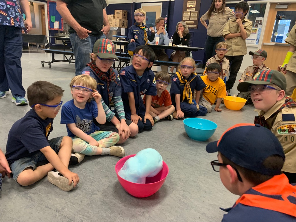 Young elementary-aged students wear safety goggles and watch as blue foam builds out of a pink bowl in the middle of a school room