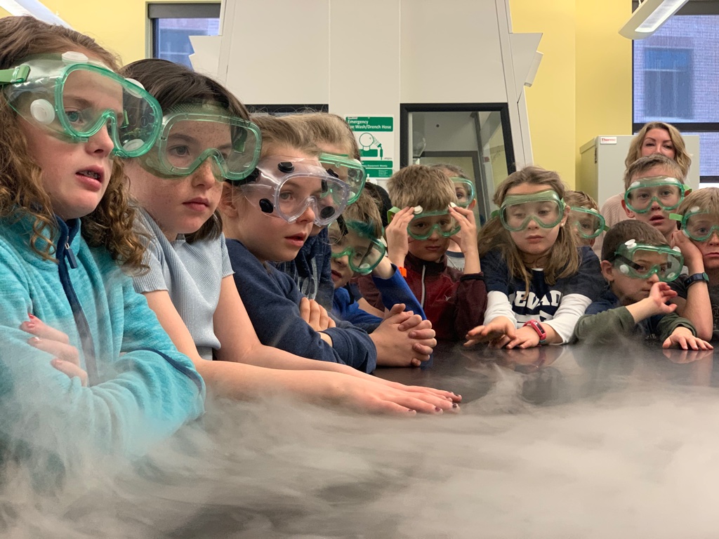 Elementary-aged students wearing green and clear safety goggles watch a chemistry demonstration. White fog is moving across the table in front of them.
