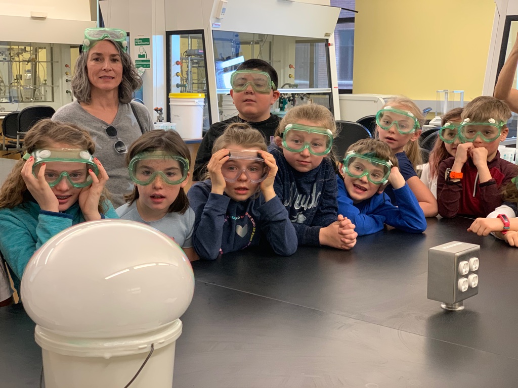 Elementary-aged students wear safety goggles and watch in awe as a chemistry demonstration is performed