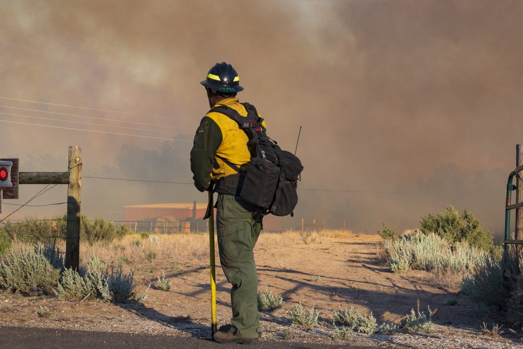 A firefighters stands in the foreground looking off into the distance at a smoke-filled background in a desert landscape