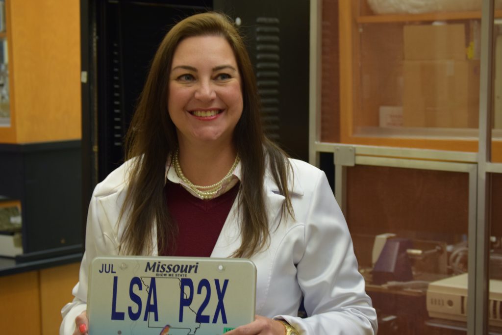 A portrait photo of Dr. Antonio, who is wearing a lab coat. She holds up a custom Missouri-issued license plate, which reads: LSA P2X.