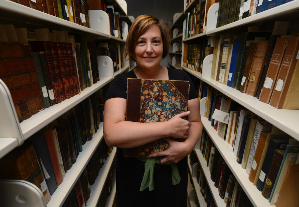 Author Jessy Randall stands in between bookshelves in library holding a book