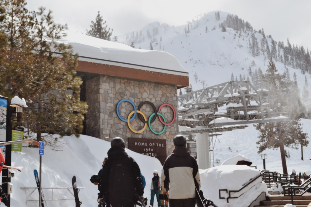 Two snowboards approach gondola with building and olympic rings in background