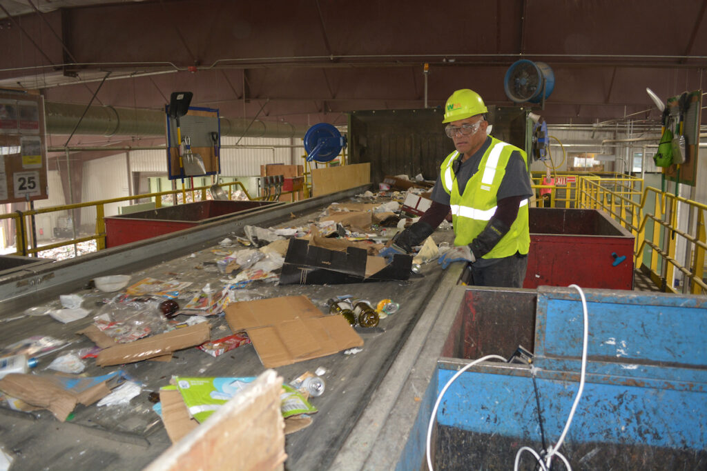 man in safety vest and helmet sorts recyclables on conveyor belt