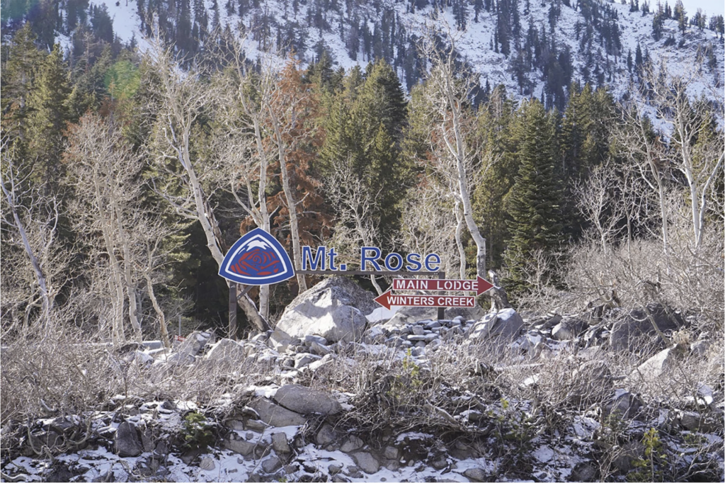 The Mt.Rose sign sits planted in the ground. Underneath, two signs in the shapes of arrows provide directions: "main lodge" to the right, and "winters creek" to the left. The signage is surrounded by rocks and snow in the foreground, and a thick forest in the background.