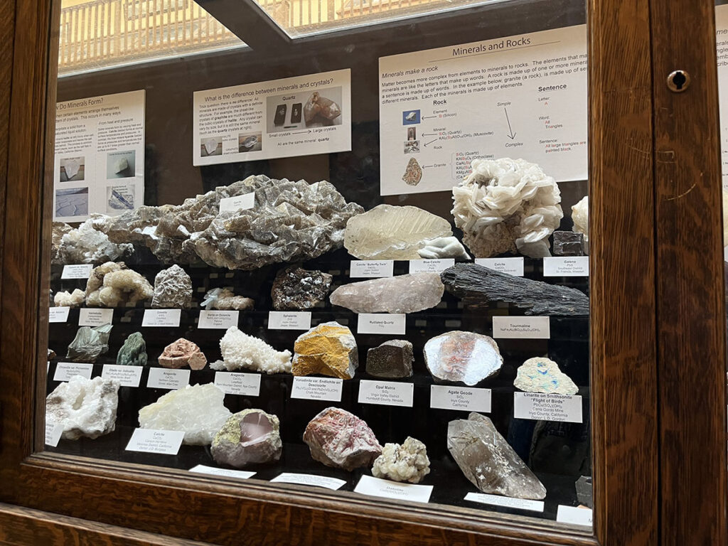 Four rows of minerals of all shapes, sizes, and colors sit in a display in the Keck Earth Science and Mineral Engineering Museum. The minerals are kept in a glass display case, and names of the minerals are provided as well as explanatory signs titles "how do minerals form?", "what is the difference between minerals and crystals", and "minerals and rocks".