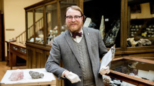 Garrett Barmore, curator of UNR’s Keck Earth Science and Mineral Engineering Museum, smiles for a picture while holding a clipboard and leaning on a tray holding two minerals.
