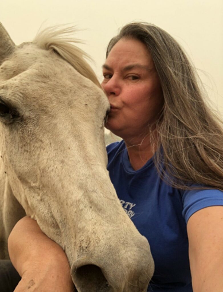 A woman embraces and kisses a horse on the head against a smoky sky